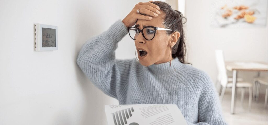 Woman looking shocked at a thermostat and holding a bill, reacting to high energy costs.